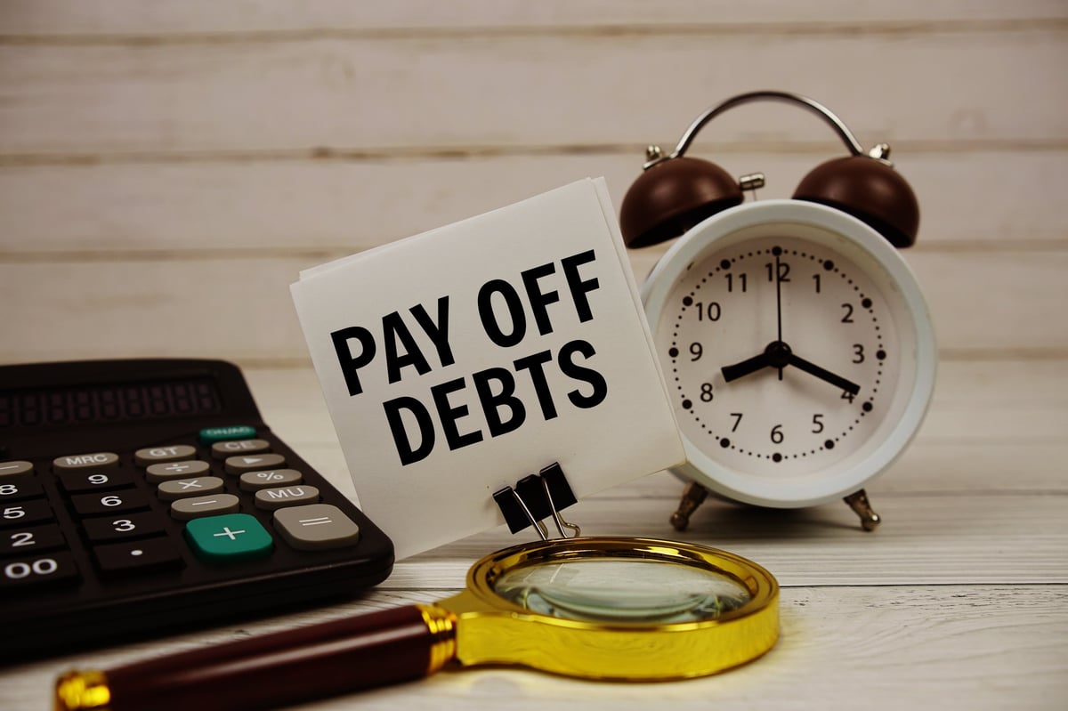 Pay Off Debts text on paper card with alarm clock, calculator and magnifying glass on wooden background
